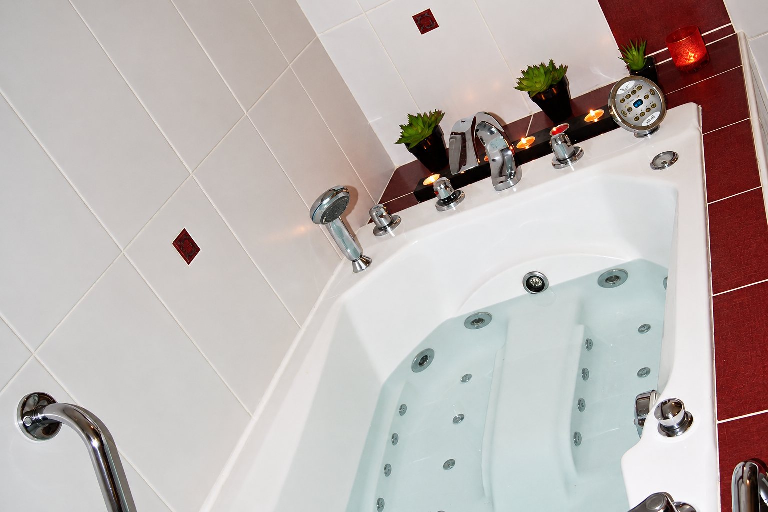 Une baignoire balnéo blanche avec jets et robinetterie chromée, décorée de bougies et de plantes grasses, entourée de carrelage blanc et bordeaux.