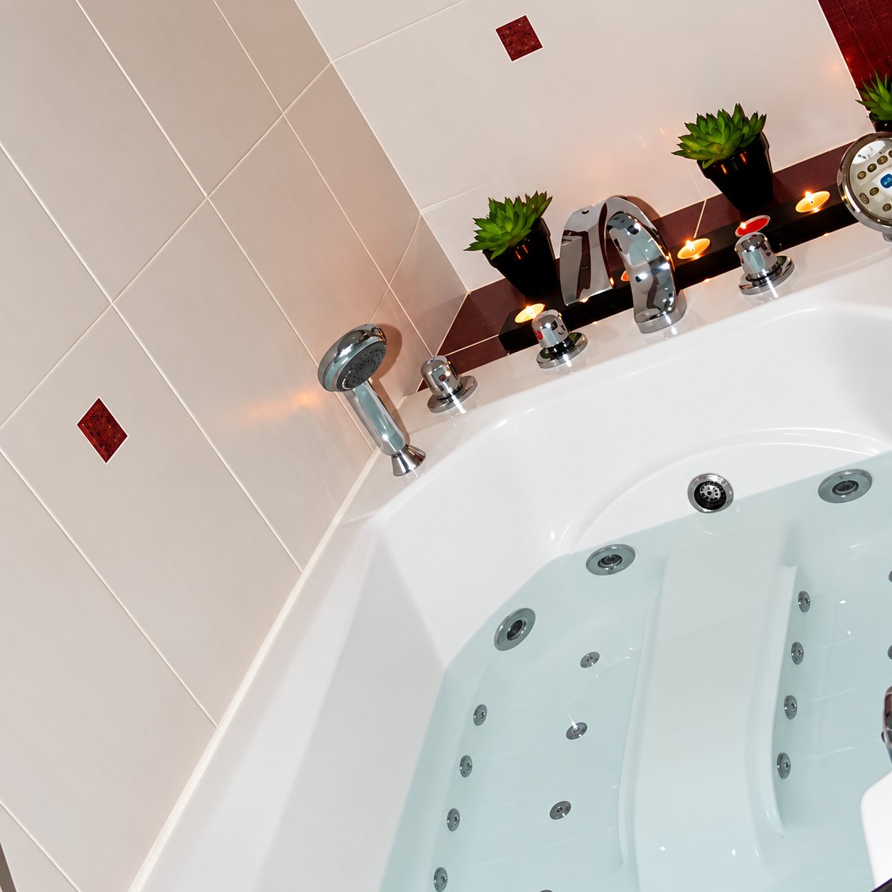 Baignoire balnéo à jets avec robinetterie chromée, petites plantes et bougies, entourée de carrelage beige orné de motifs rouges.