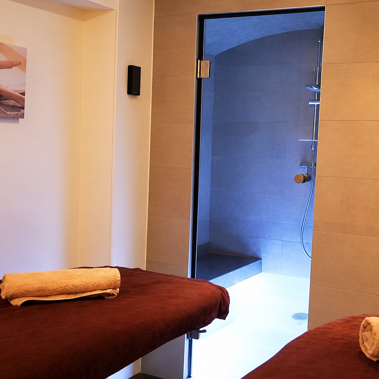 Salle de soins avec deux tables de massage couvertes de draps bordeaux et de serviettes, devant un hammam à porte vitrée éclairé en bleu.