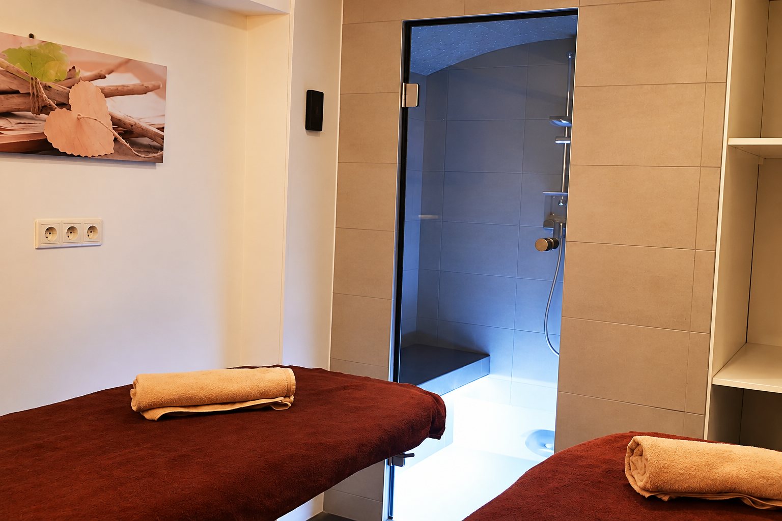 Une cabine de soins avec deux tables de massage bordeaux ornées de serviettes roulées, face à un hammam éclairé derrière une porte en verre.
