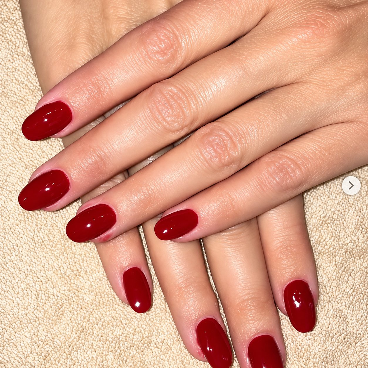 Deux mains posées l'une sur l'autre sur une serviette beige, avec des ongles ovales vernis en rouge foncé brillant.