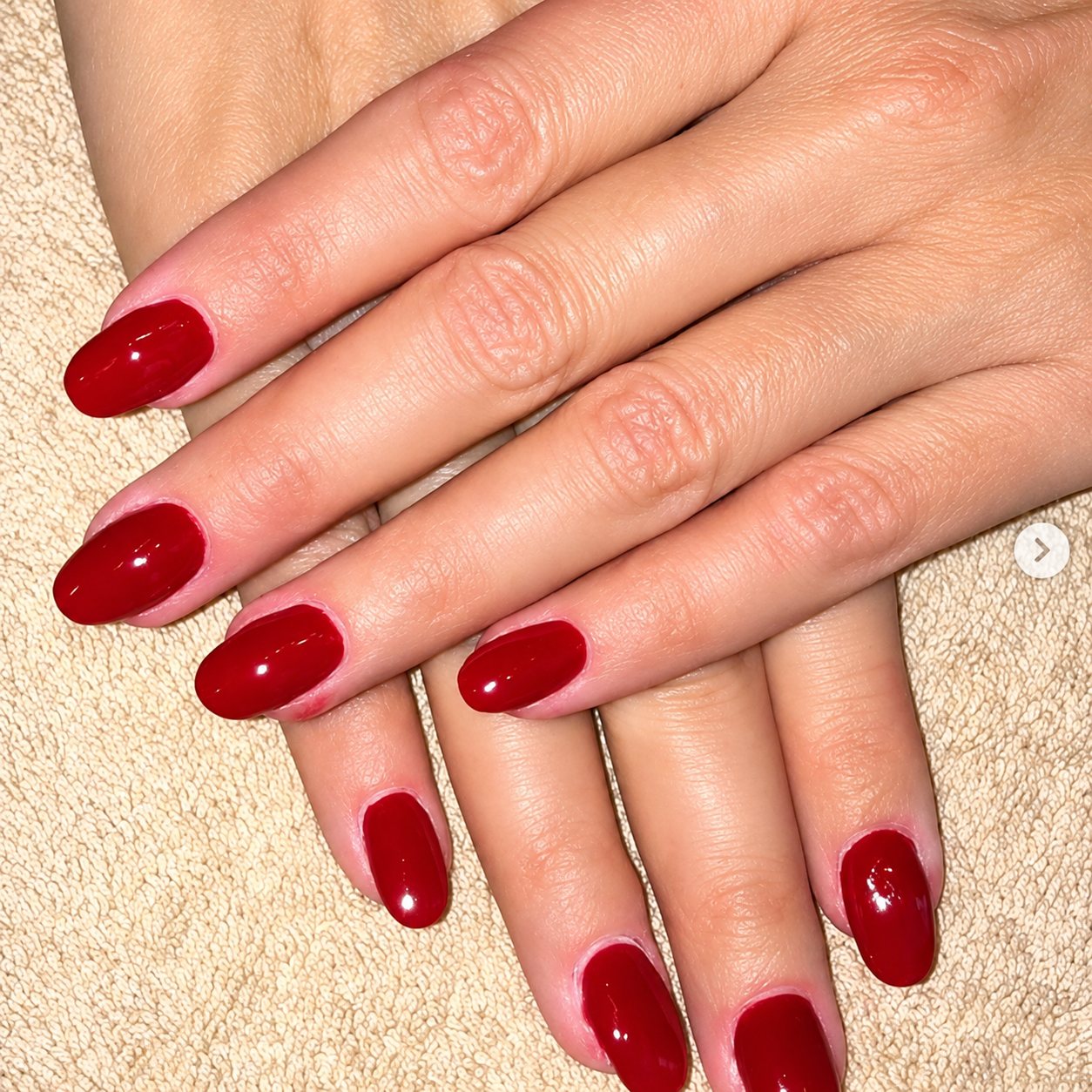 Deux mains posées sur une serviette beige montrant des ongles ovales recouverts d'un vernis rouge foncé brillant.