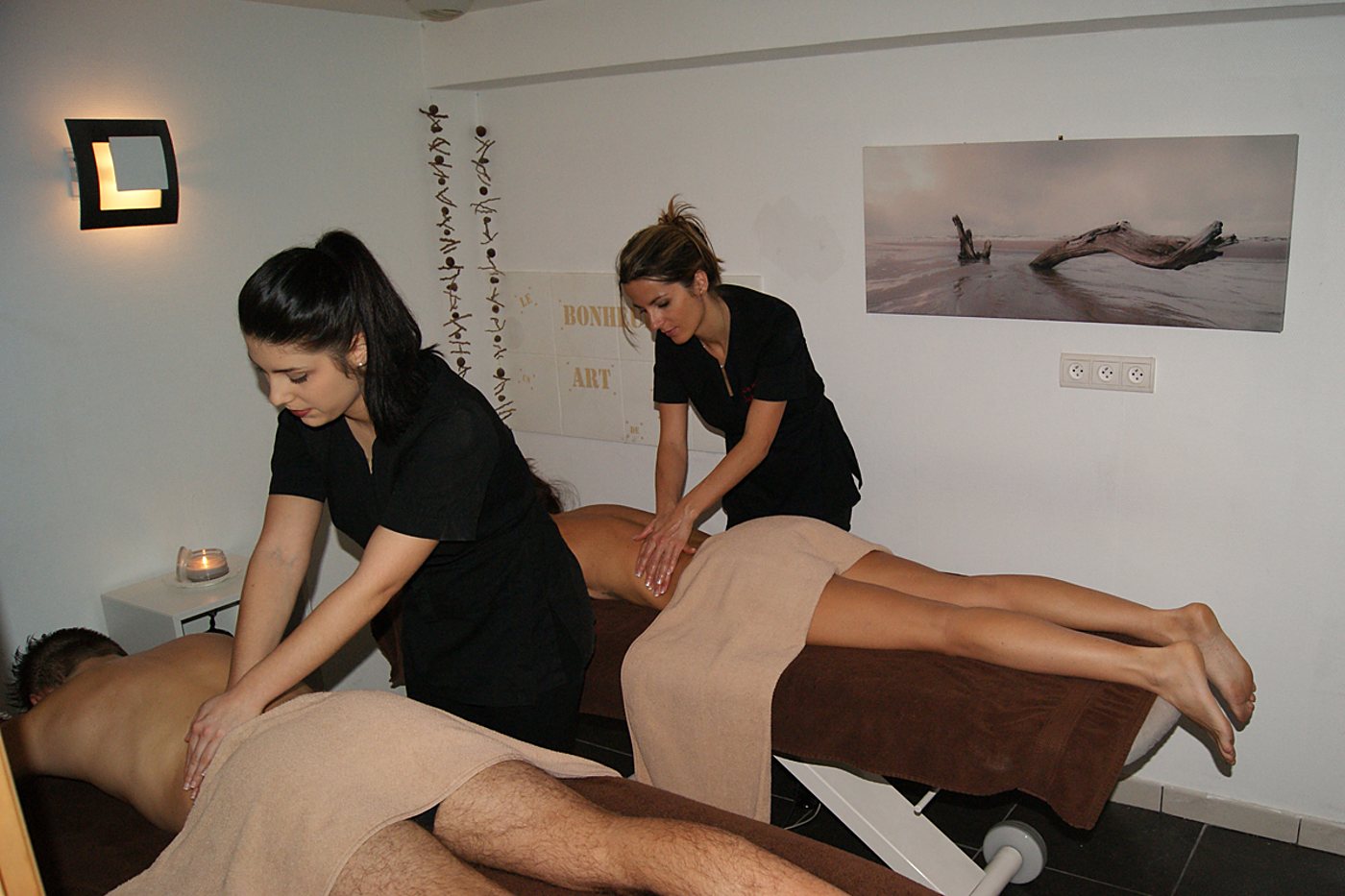 Deux praticiennes en tenue noire massent le dos de deux clients allongés sur des tables de massage dans une cabine claire avec bougie et tableau de bois flotté.