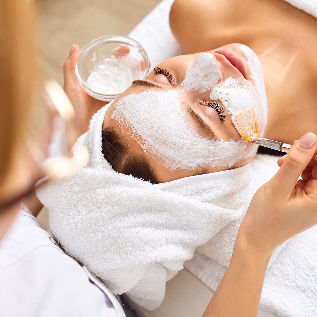 Une esthéticienne applique au pinceau un masque blanc sur le visage d'une femme allongée, les cheveux enveloppés dans une serviette blanche.