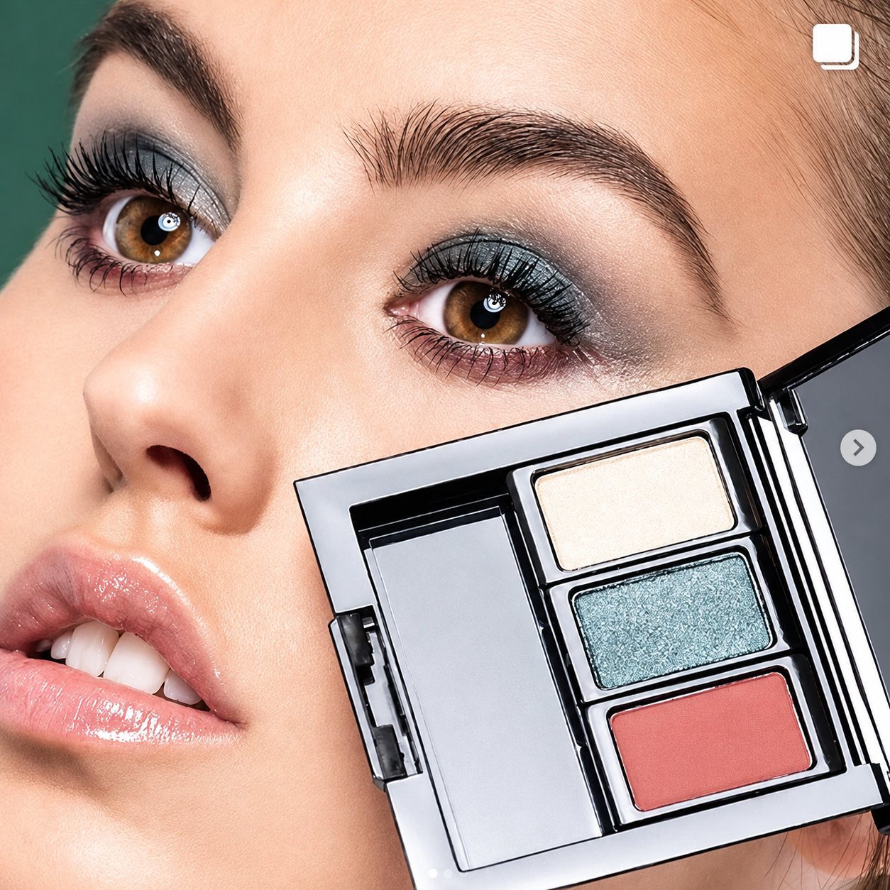 Gros plan sur le visage d'une femme aux yeux maquillés de gris-vert, tenant près de sa joue une palette de trois fards à paupières dans un boîtier argenté.